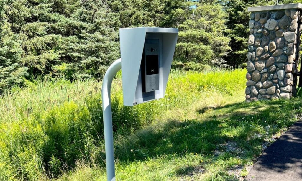 An outdoor intercom system stands at the entrance to a property, featuring a gray metal housing and a black screen, against a backdrop of lush green foliage and a stone pillar.