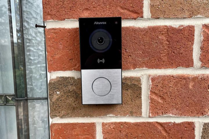 Akuvox smart doorbell mounted on a red brick wall next to a leaded glass window. The doorbell has a black top section with camera and wifi symbol, and a silver bottom section with a circular button.