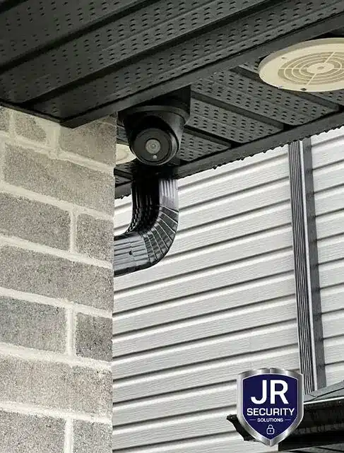 Dome camera mounted under a dark porch roof beside grey brick and siding, one of the security systems we install